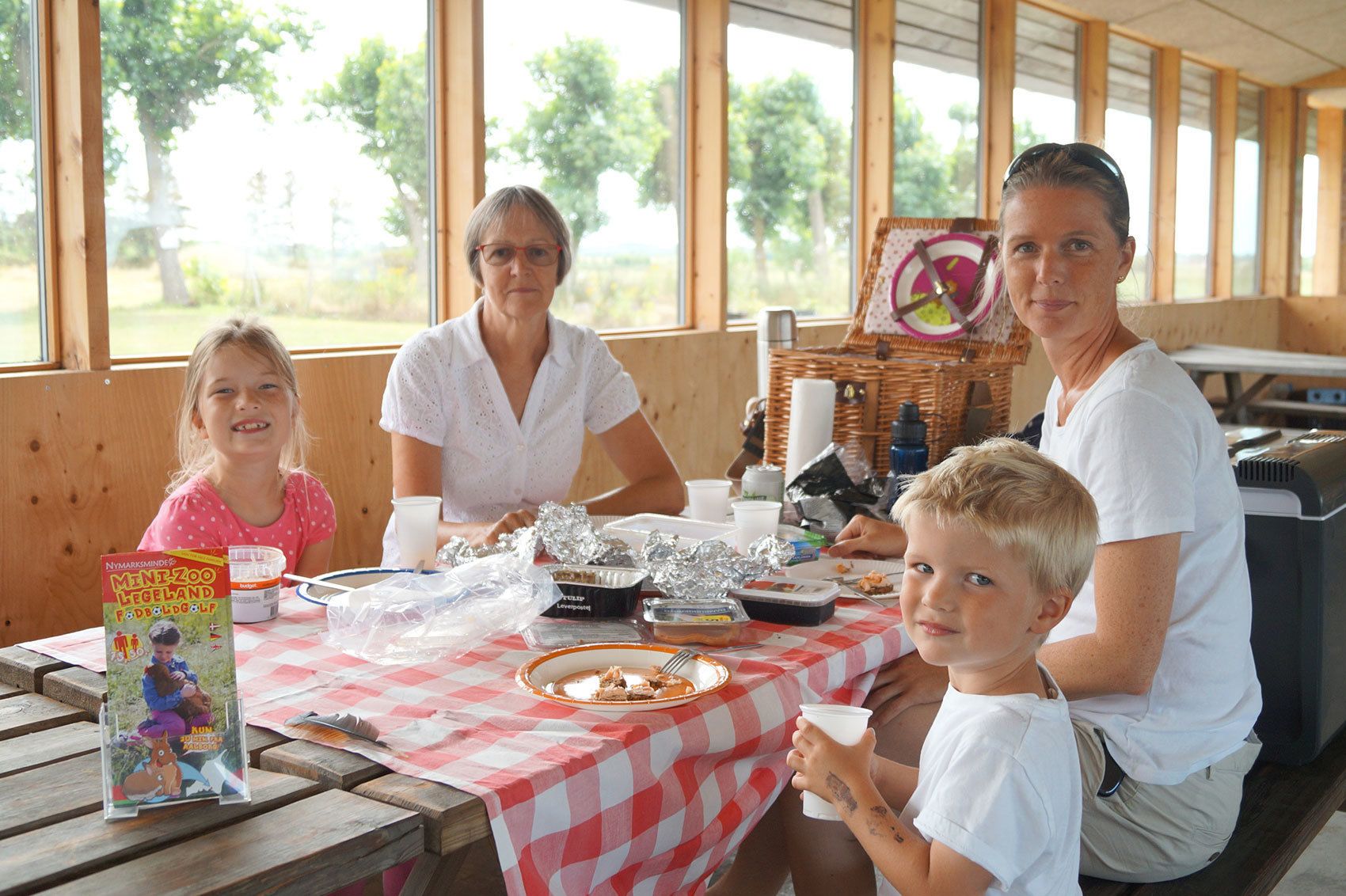 A family dining at a wooden table with a checkered tablecloth, surrounded by windows with a view of trees.