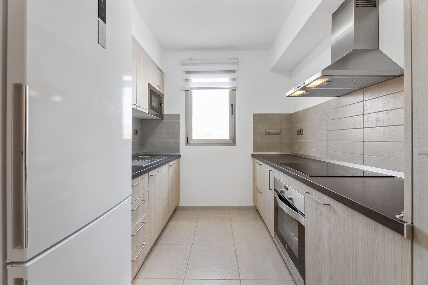 A narrow kitchen with white cabinets, a stainless steel range hood, and a window above the sink.