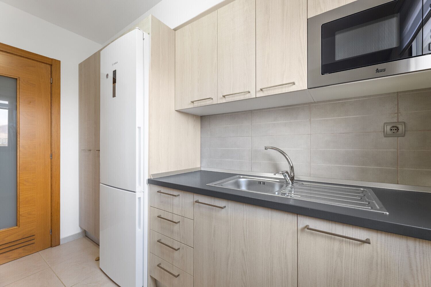 A modern kitchen with light wood cabinets, a stainless steel sink, and a built-in microwave.