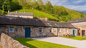 A stone building with a gravel courtyard, a dining area with outdoor seating, and a view of a green hillside. - Heron Cottage, Upperdale | Peak District (Buxton)