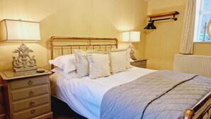 A neatly made bed with white linens and a blue quilt, a wooden nightstand with a lamp, and a wall-mounted shelf with a lamp. - Heron Cottage, Upperdale | Peak District (Buxton)
