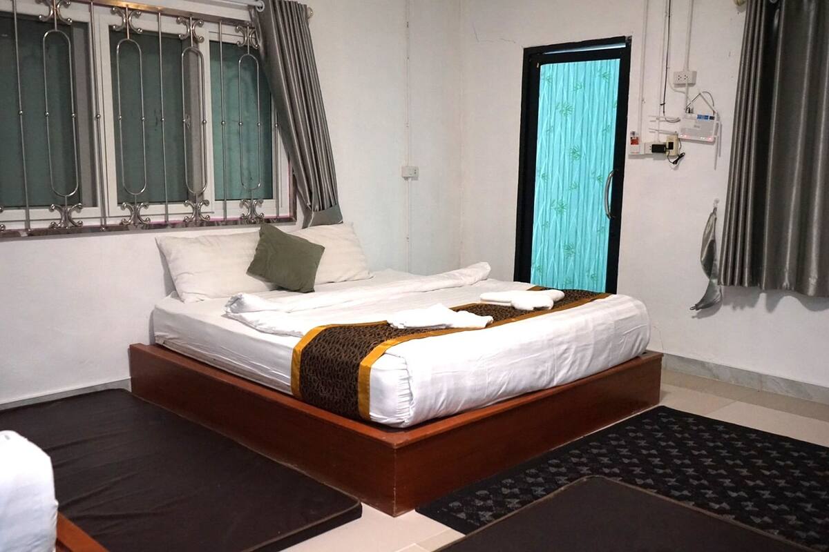 A neatly made bed with white linens and a brown accent, a window with metal bars, a door with a curtain, and a wall-mounted power outlet.