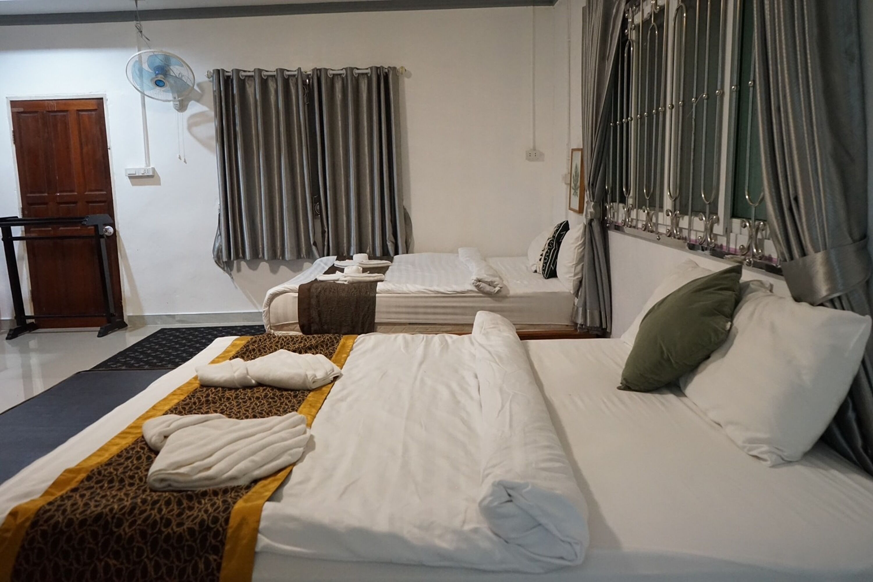 A hotel room with a bed, a desk, a chair, a standing fan, and curtains.