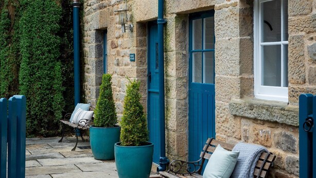 A stone building with a blue door and window, a bench with cushions, and potted plants. - Stable Cottage Chatsworth Estate (Baslow)