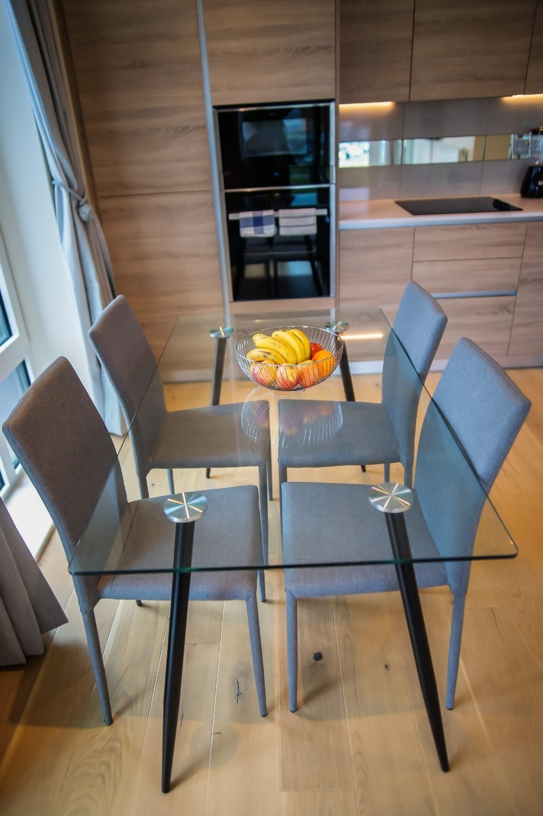 Una cucina moderna con un tavolo da pranzo in vetro e sedie, una ciotola di frutta sul tavolo e mobili in legno.