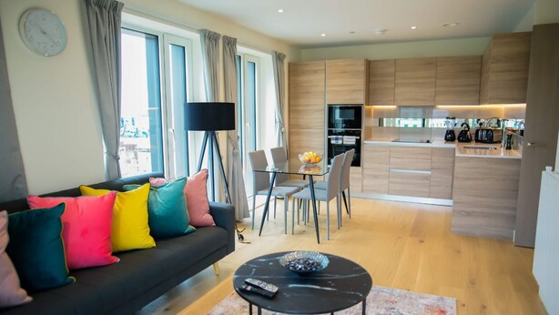 A modern living room with a black sofa, colorful cushions, a glass coffee table, and a dining area with a table and chairs. - Thames View 2 Bed Apartment With Balcony (London)