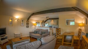 A cozy living room with a sofa, a coffee table, and a TV. There's a kitchen area with a dining table and chairs. The room has wooden beams on the ceiling and wall-mounted lights. - Heathy Lea Barn Chatsworth Estate (Baslow)