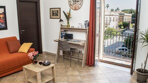 Uma sala de estar com sofá, escrivaninha e vista para a rua através de uma porta de varanda.