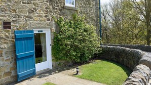 Un bâtiment en pierre avec une porte blanche et des volets bleus, une petite pelouse et un mur en pierre.