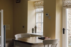 A kitchen with a wooden table, white chairs, a window with floral curtains, and a stone wall.
