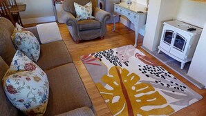 A cozy living room with a sofa, armchair, and a patterned rug. A kitchen area is visible in the background. - Rowsley Cottage, Bakewell | Peak District (Bakewell)