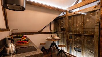 A rustic kitchen with a wooden beam ceiling, a gas stove, a kettle, and a wooden table with stools.