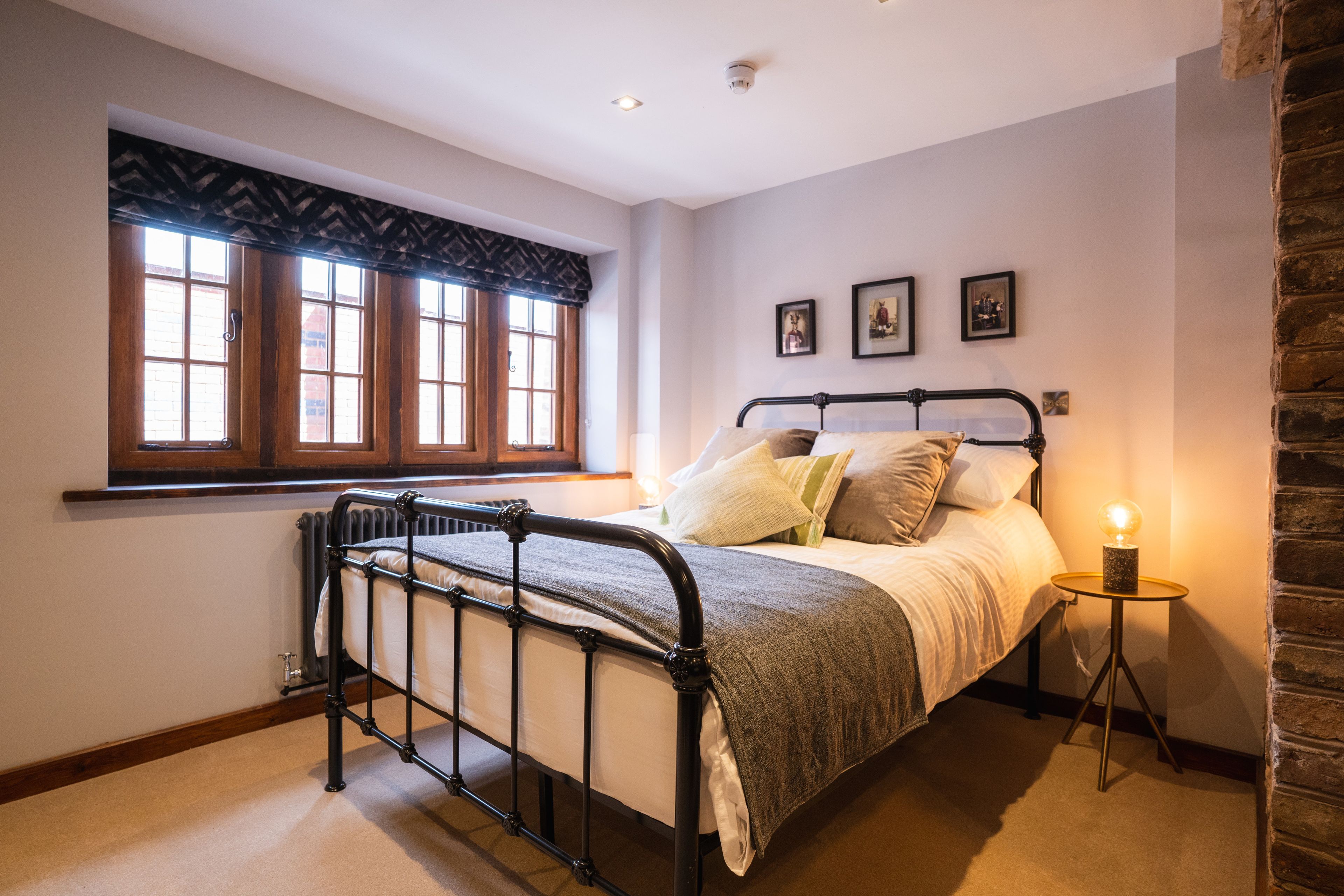 A bedroom with a bed, a window with curtains, a bedside table with a lamp, and a brick wall.