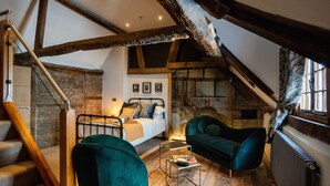 Un dormitorio tipo loft con vigas de madera a la vista, una cama con almohadas, un sillón verde, una mesita y una escalera.