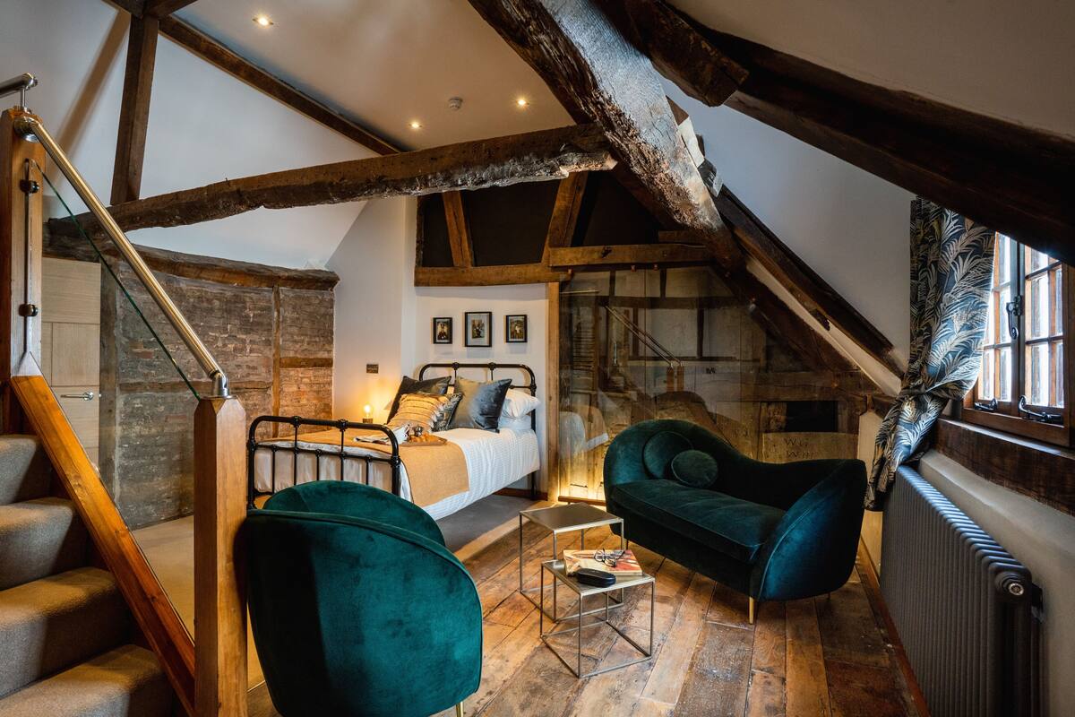 A loft bedroom with exposed wooden beams, a bed with pillows, a green armchair, a small table, and a staircase.