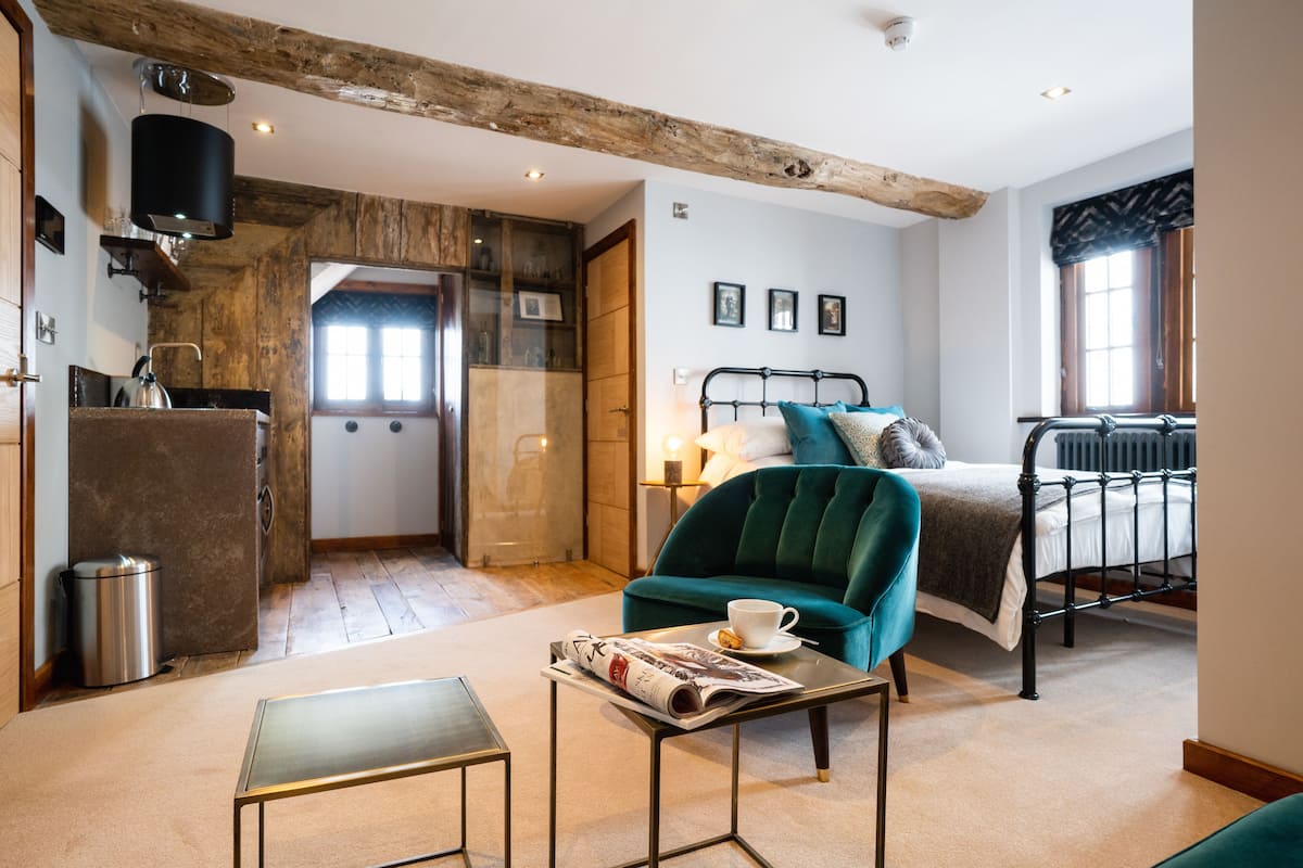A cozy bedroom with a bed, a green armchair, a small table with a newspaper, and a radiator.