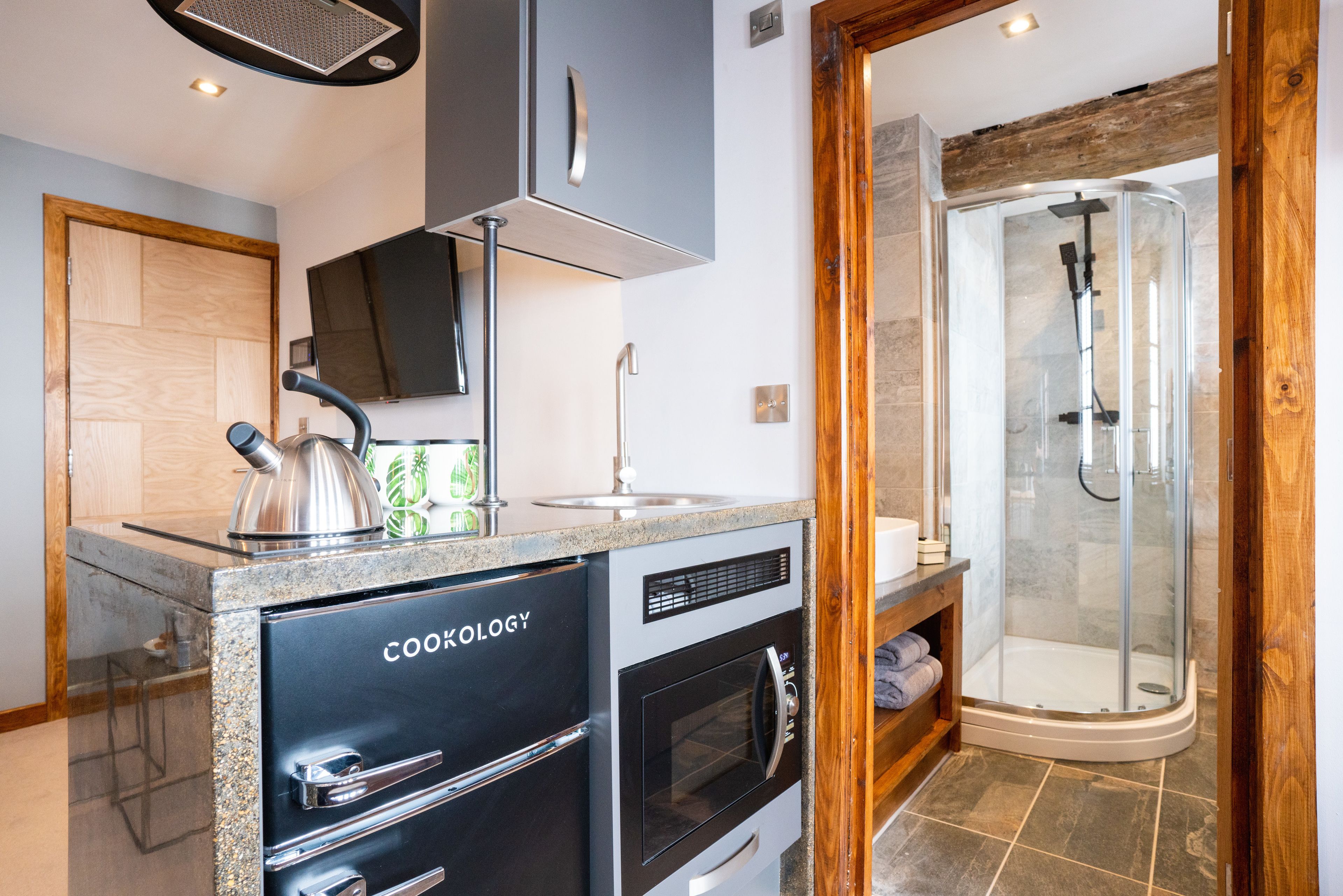 A compact kitchen with a built-in oven and a coffee machine, a glass shower enclosure, and a wooden door.