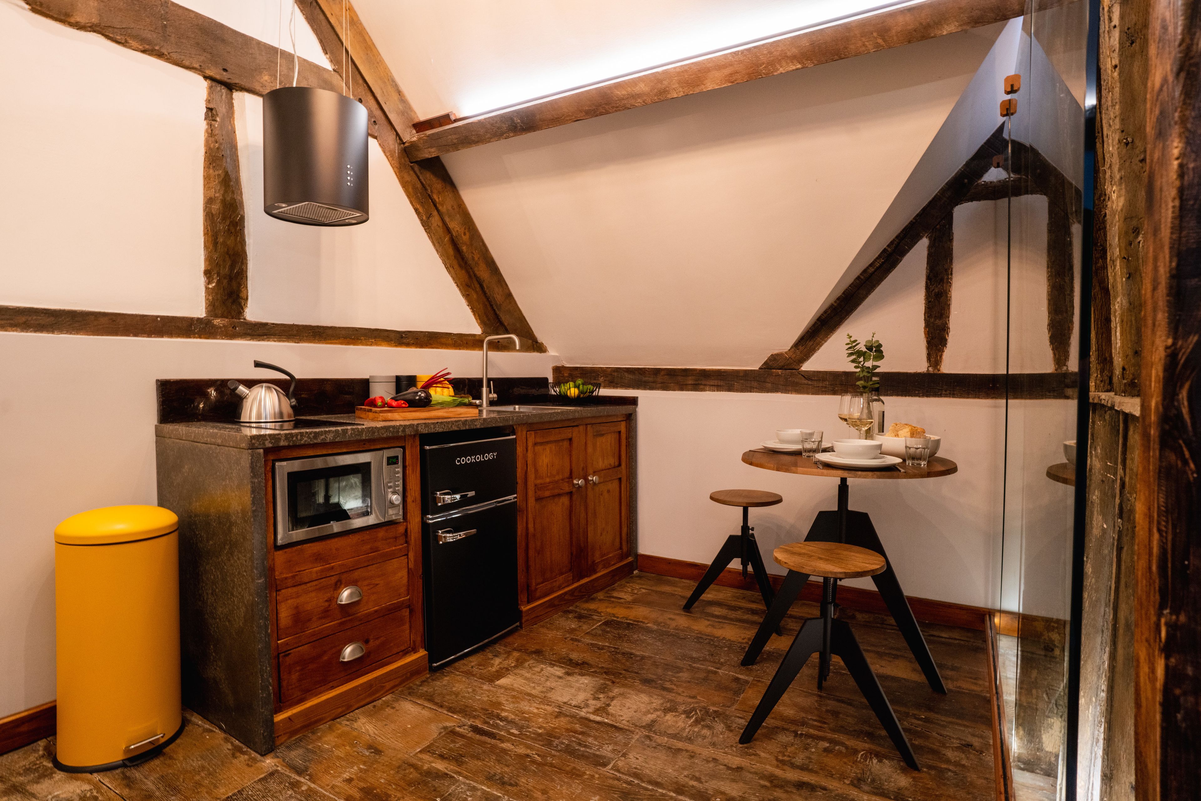 A compact kitchen with wooden beams, a built-in cooktop, a black dishwasher, and a small round table with stools.