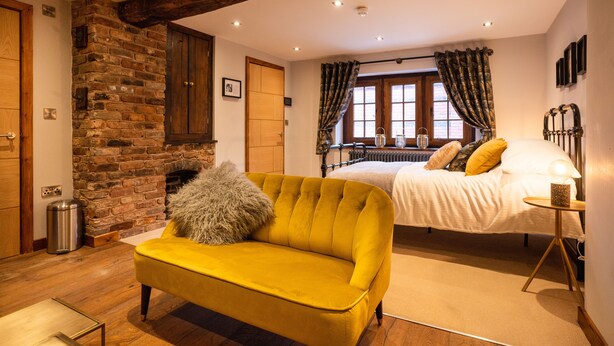 Un dormitorio con cama, un sofá amarillo, chimenea de ladrillo y un travesaño de madera.
