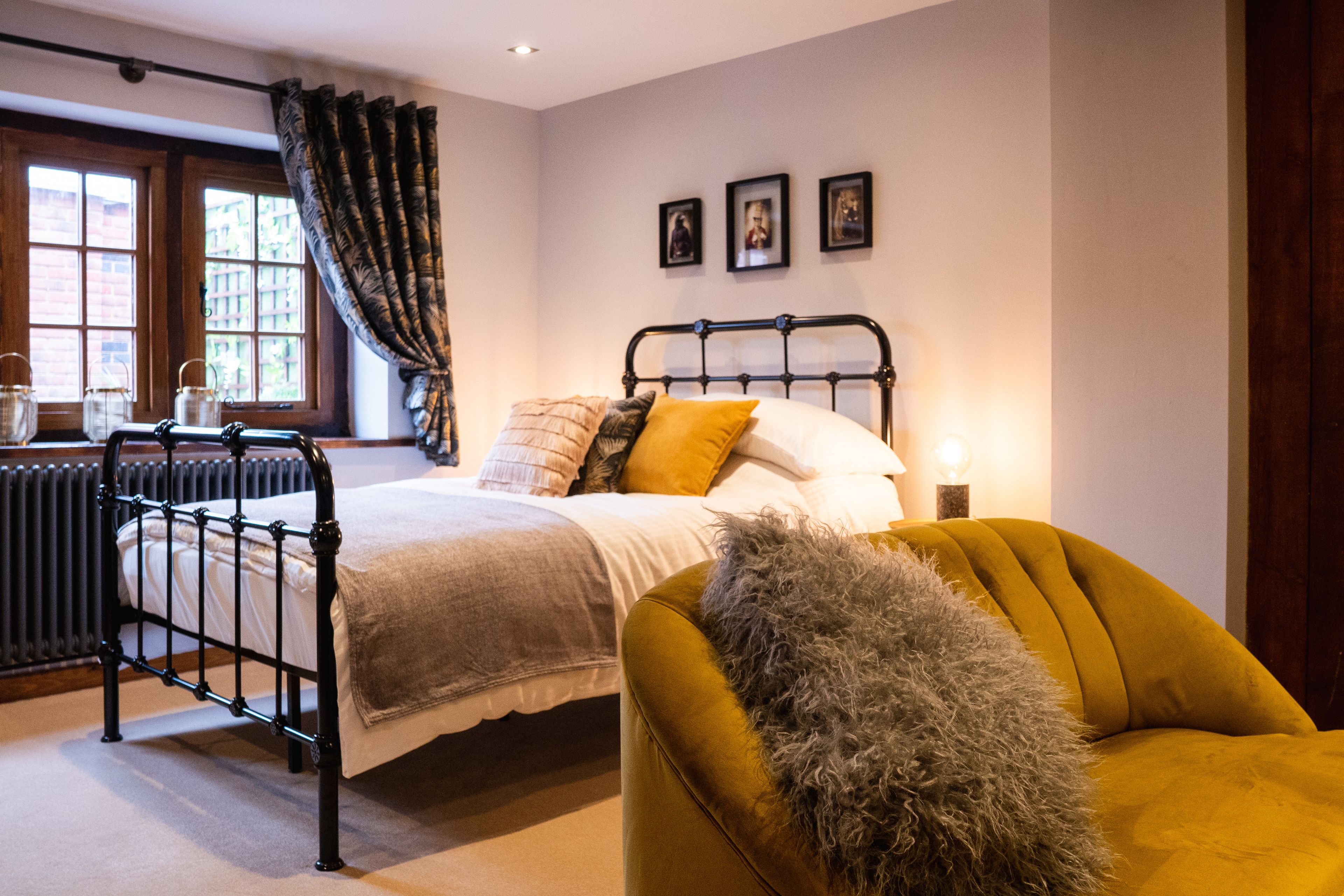A bedroom with a bed, a chair, a radiator, a window with curtains, and framed pictures on the wall.