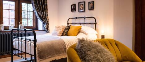 A bedroom with a bed, a chair, a radiator, a window with curtains, and framed pictures on the wall.