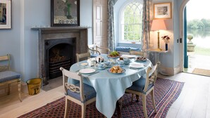 A dining room with a round table set for breakfast, a fireplace, and a view of a lake through a window. - Swiss Cottage Chatsworth Estate (Chatsworth)