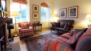 A cozy living room with a plaid armchair, a sofa, and a patterned rug. There are two lamps, a fireplace, and framed pictures on the wall. - Russian Cottage Chatsworth Estate (Edensor)