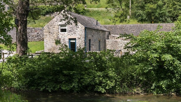 A stone building with a blue door, surrounded by lush greenery and a river. - Dipper Cottage Upperdale | Peak District (Buxton)
