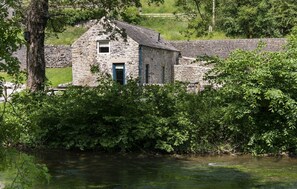 A stone building with a blue door, surrounded by lush greenery and a river.