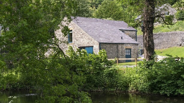 A stone building with a sloped roof, surrounded by lush greenery and a body of water. - Kingfisher Cottage Upperdale | Peak District (Buxton)