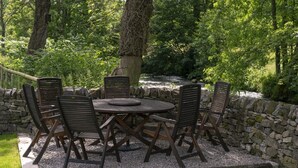 An outdoor seating area with a round table and six chairs, surrounded by a stone wall and lush greenery. - Kingfisher Cottage Upperdale | Peak District (Buxton)