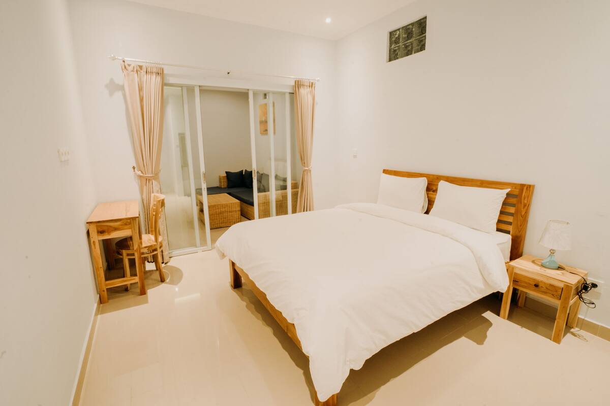 A bedroom with a wooden bed, a nightstand, a small table, and a chair. There is a sliding glass door leading to a balcony.