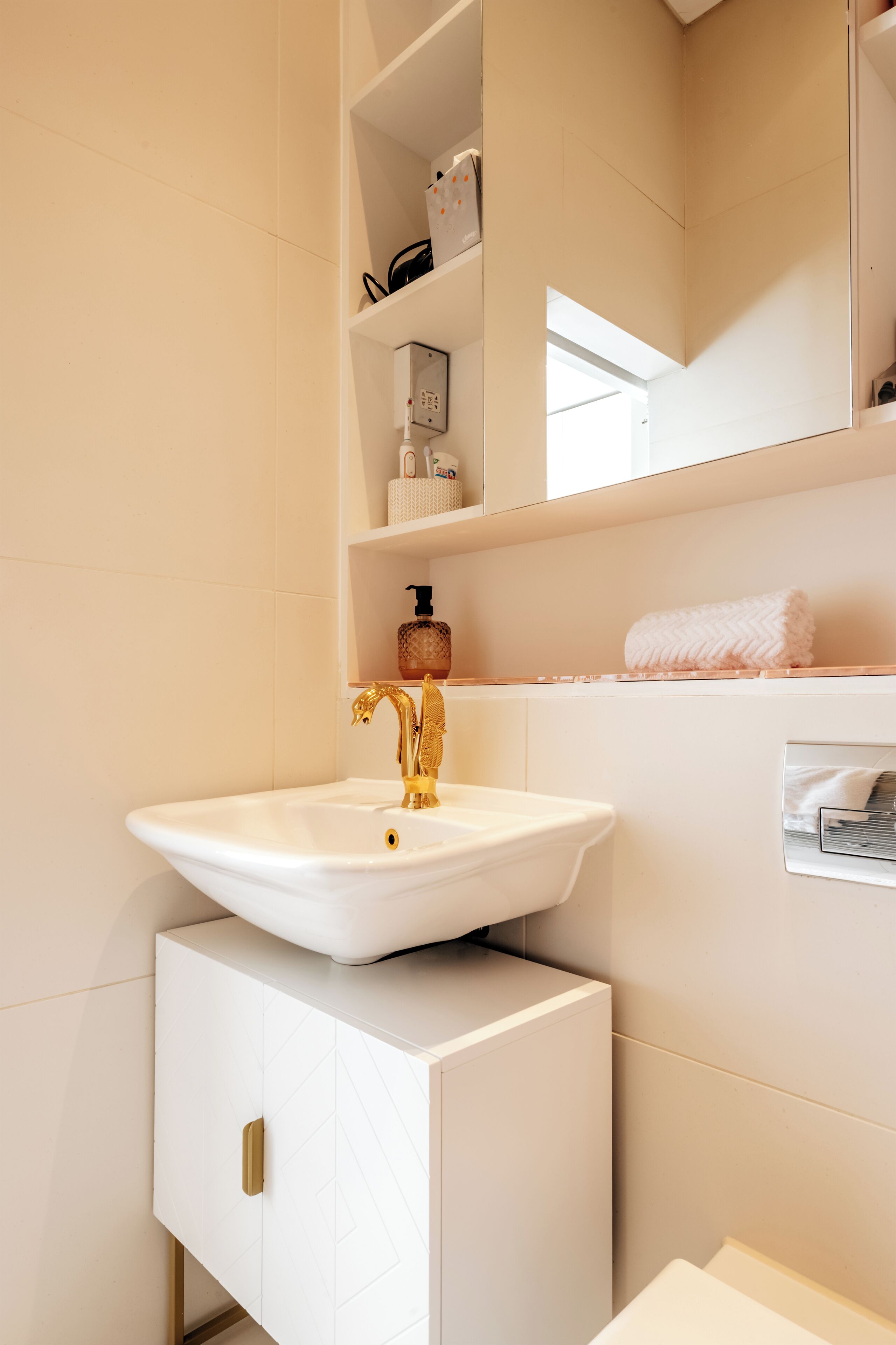 Un bagno con un lavandino bianco, un rubinetto dorato e un mobile bianco con maniglia dorata. Sopra il lavandino c'è uno scaffale con articoli da toilette e un asciugamano appeso al muro.