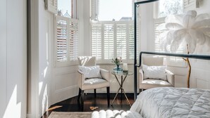 A bedroom with a bed, two chairs, a small table, and a window with shutters.