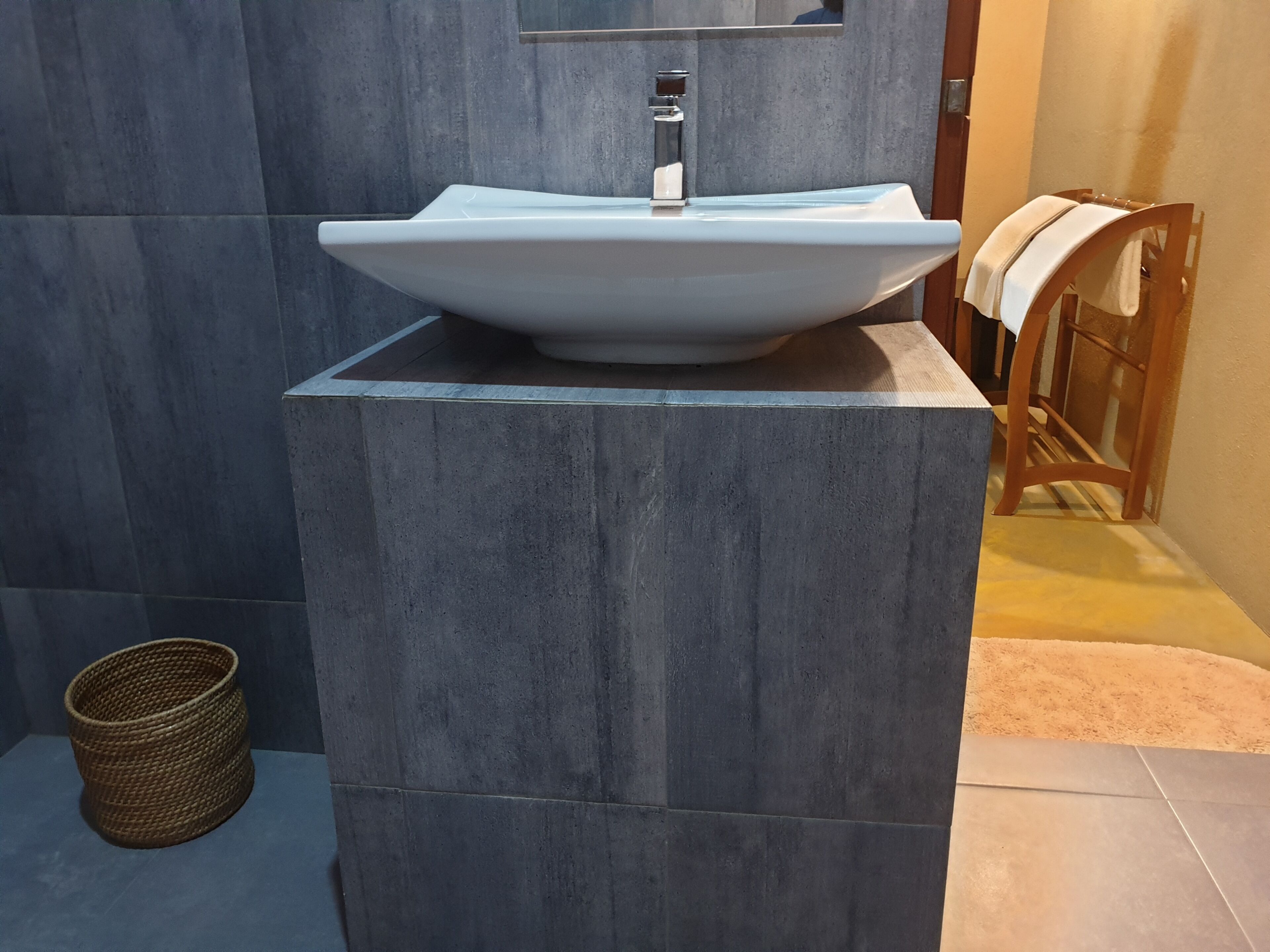 Ein Badezimmer mit einem großen, modernen Waschtisch auf einem Sockel, einem Holzstuhl mit Handtuch und einem gewebten Korb am Boden.
