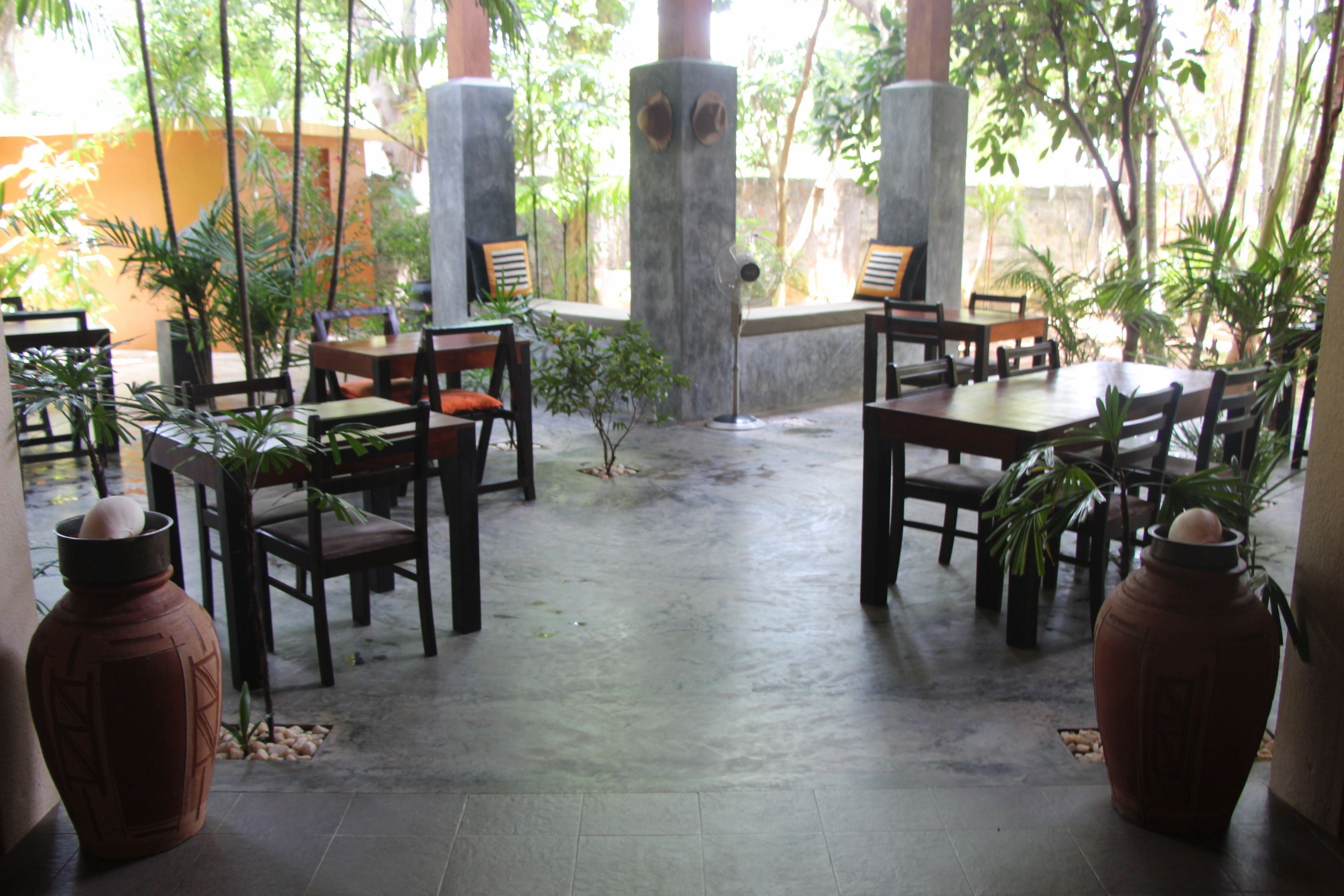 An outdoor patio with wooden tables and chairs, surrounded by potted plants and a concrete floor.