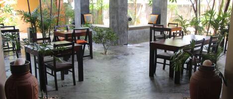 An outdoor patio with wooden tables and chairs, surrounded by potted plants and a concrete floor.