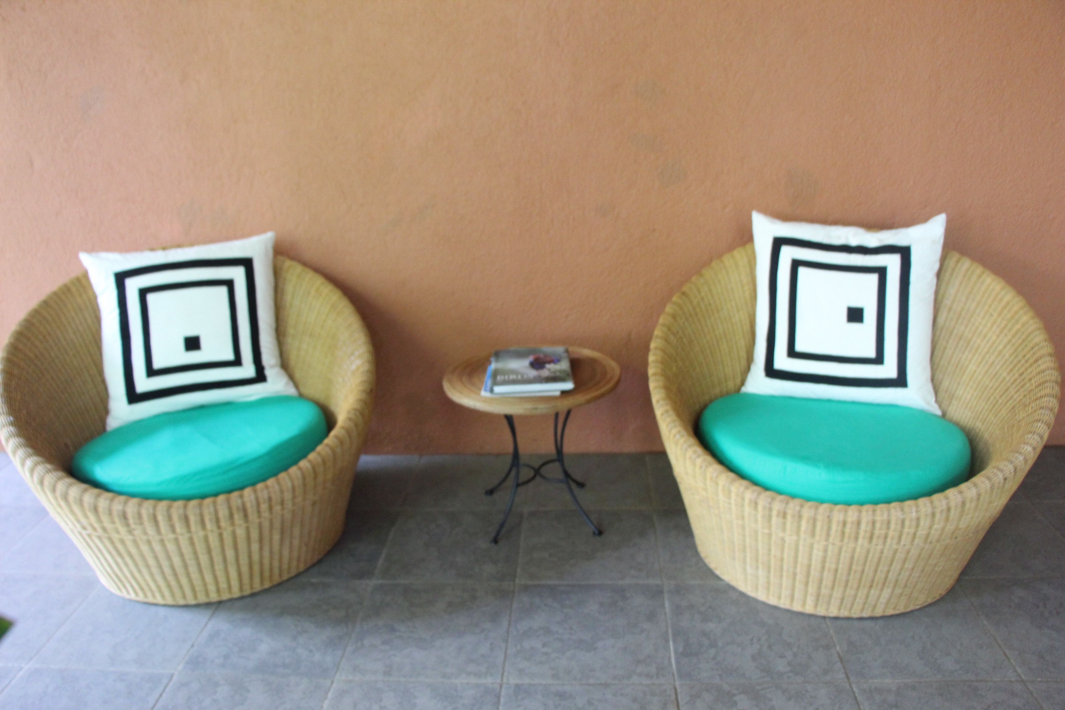 Two wicker chairs with teal cushions and black and white patterned pillows, positioned on a tiled floor with a small round table between them.