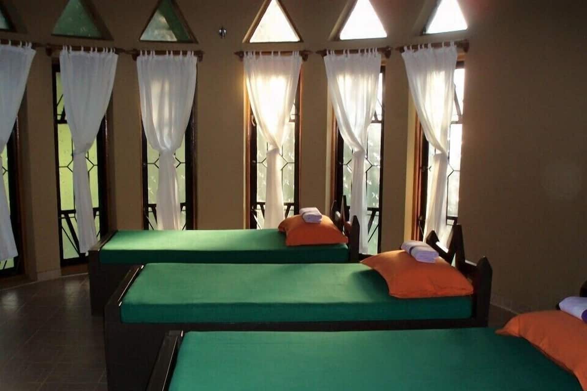 A hotel room with three single beds, each with a green bedspread and an orange pillow. The room has large windows with white curtains, allowing natural light to enter.