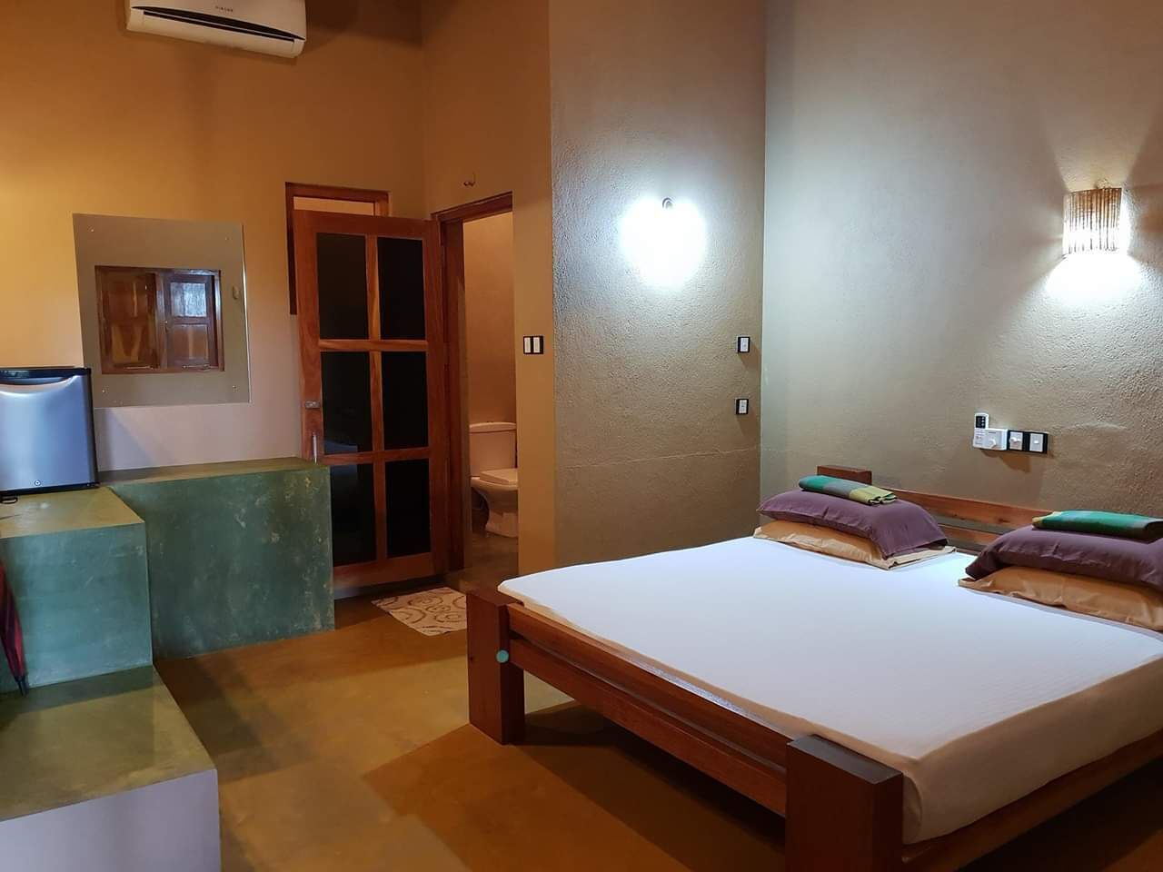 Une chambre avec un grand lit, un banc en bois, une petite table, une lampe fixée au mur et une porte donnant sur une salle de bain.