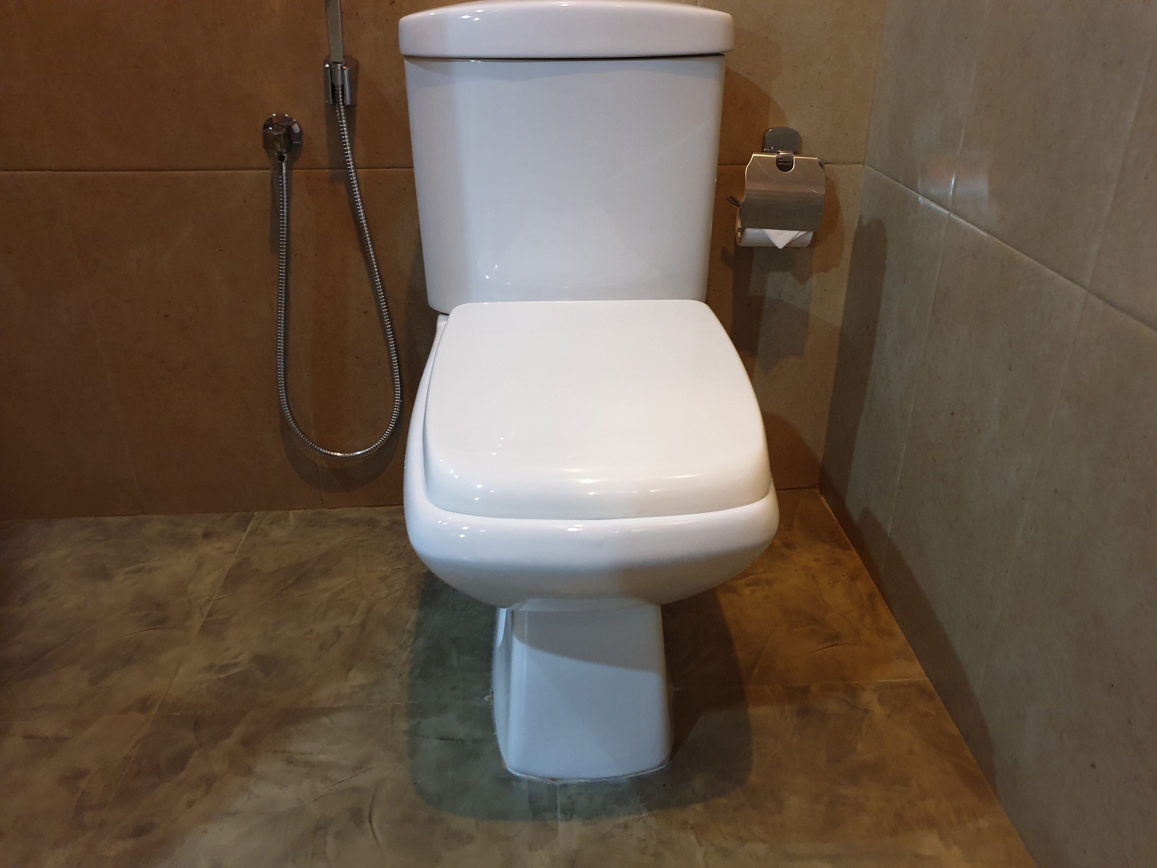 A white toilet with a shower attachment in a bathroom with brown tiled walls.