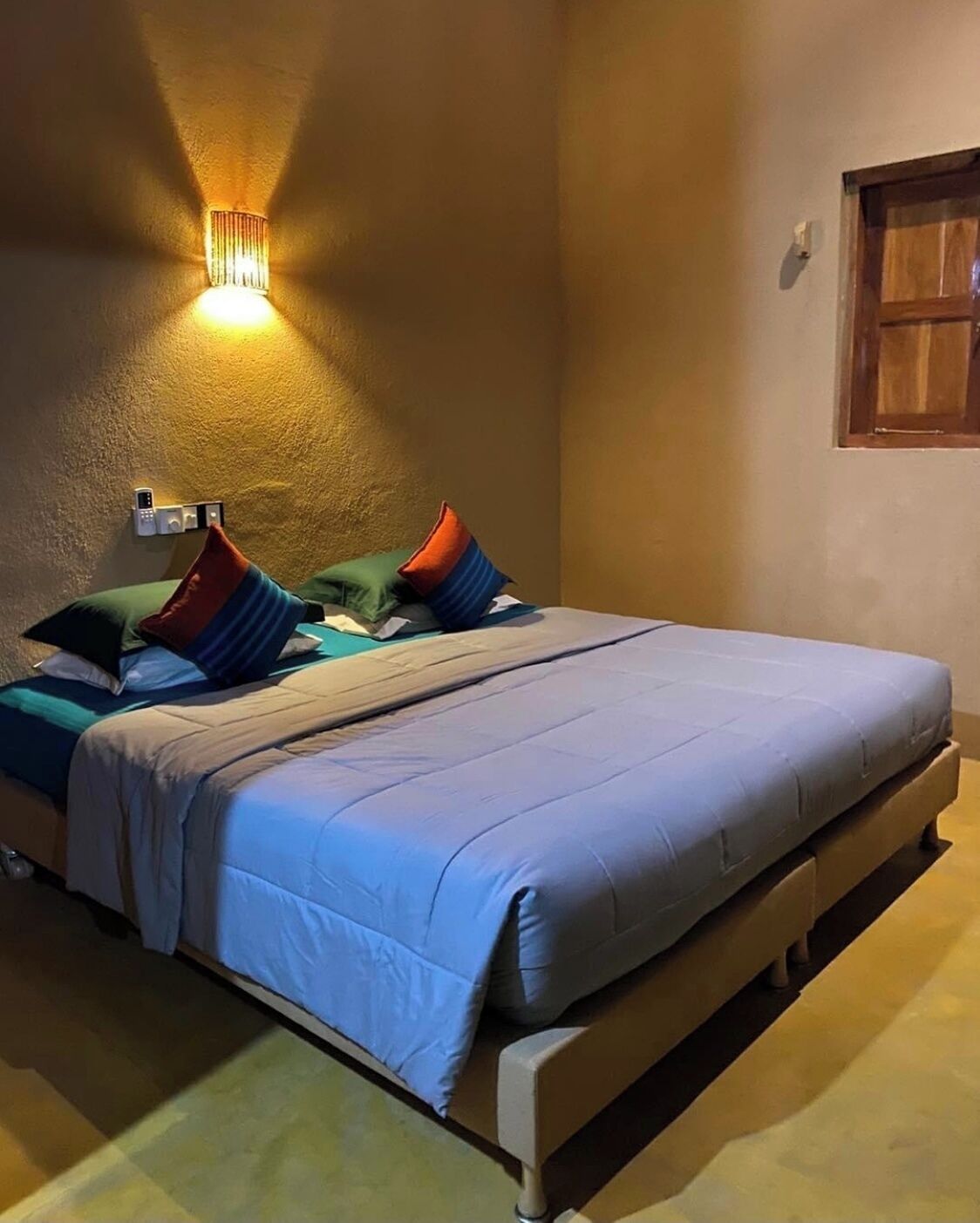 Ein ordentlich bezogenes Bett mit blauer Decke und Kissen, ein Nachttisch mit Fernbedienung und eine Wandleuchte.