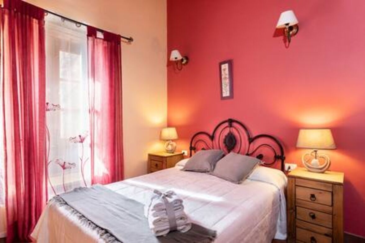 Un dormitorio con una cama, dos mesitas de noche con lámparas, una ventana con cortinas y una lámpara fijada a la pared.