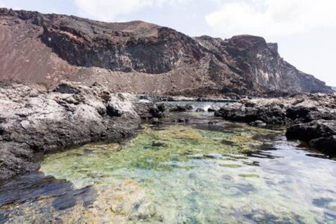 A rocky shoreline with clear water and a rugged mountain backdrop.