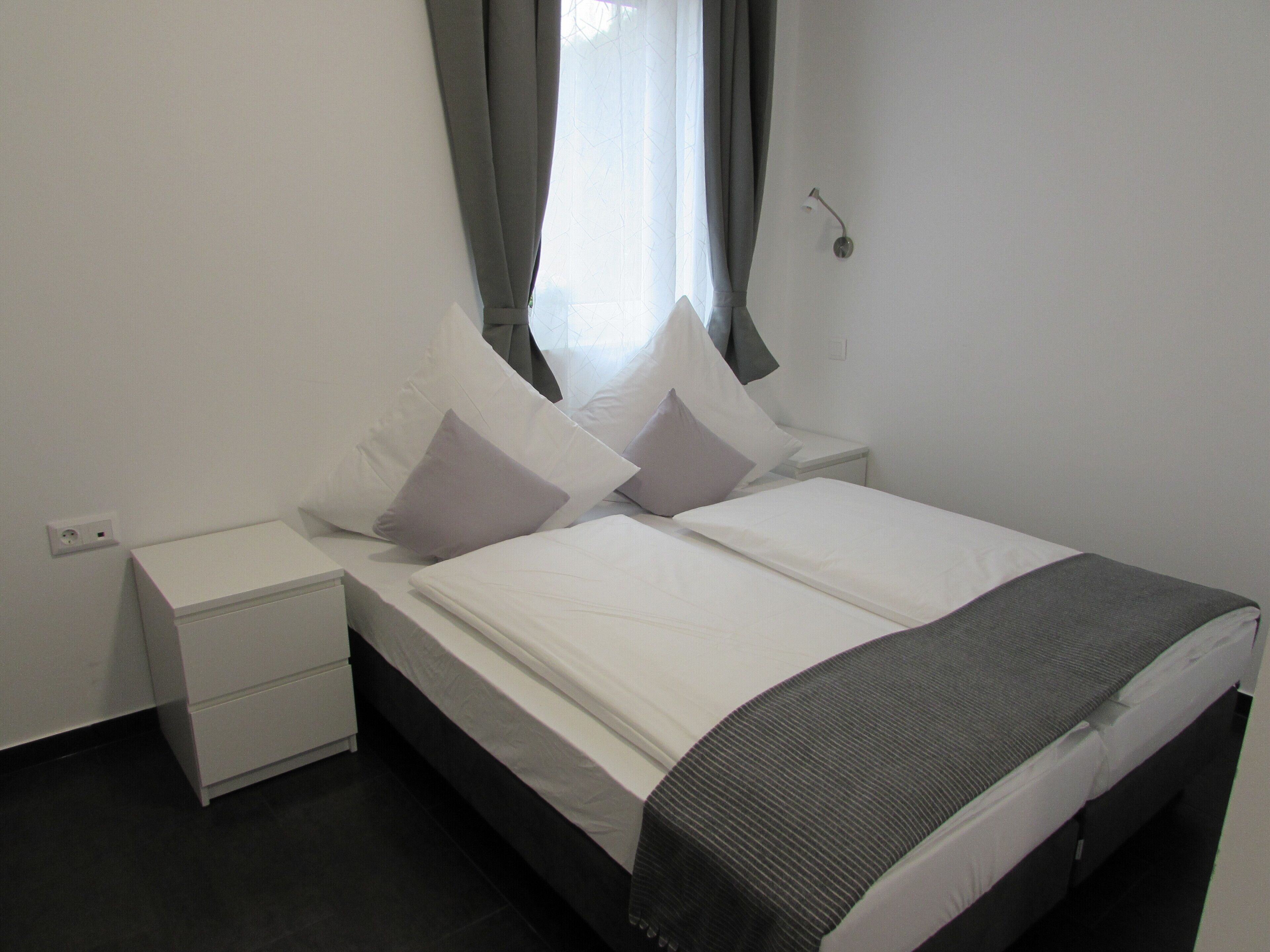 A neatly made bed with white linens and two pillows, a bedside table, and a wall-mounted light fixture.