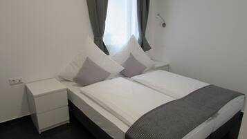 A neatly made bed with white linens and two pillows, a bedside table, and a wall-mounted light fixture.