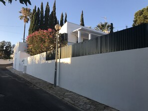 A modern white building with a tall fence, a street lamp, and a tree with pink flowers. - Luxury Beach House w / Heated Pool + Jacuzzi (Albufeira)