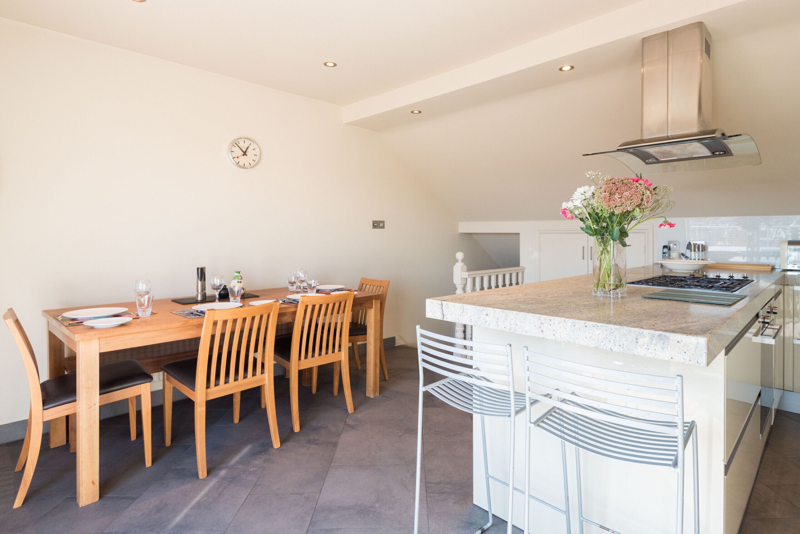 A modern kitchen with a dining area, featuring a wooden table and chairs, a built-in stove, and a clock on the wall.