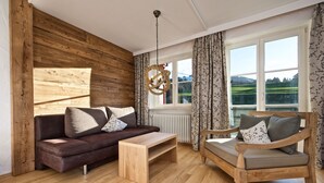 Una sala de estar acogedora con un sofá de cuero marrón, una mesa de centro de madera y un sillón de madera con cojines. Se observa un panel de madera en la pared y una ventana con cortinas.