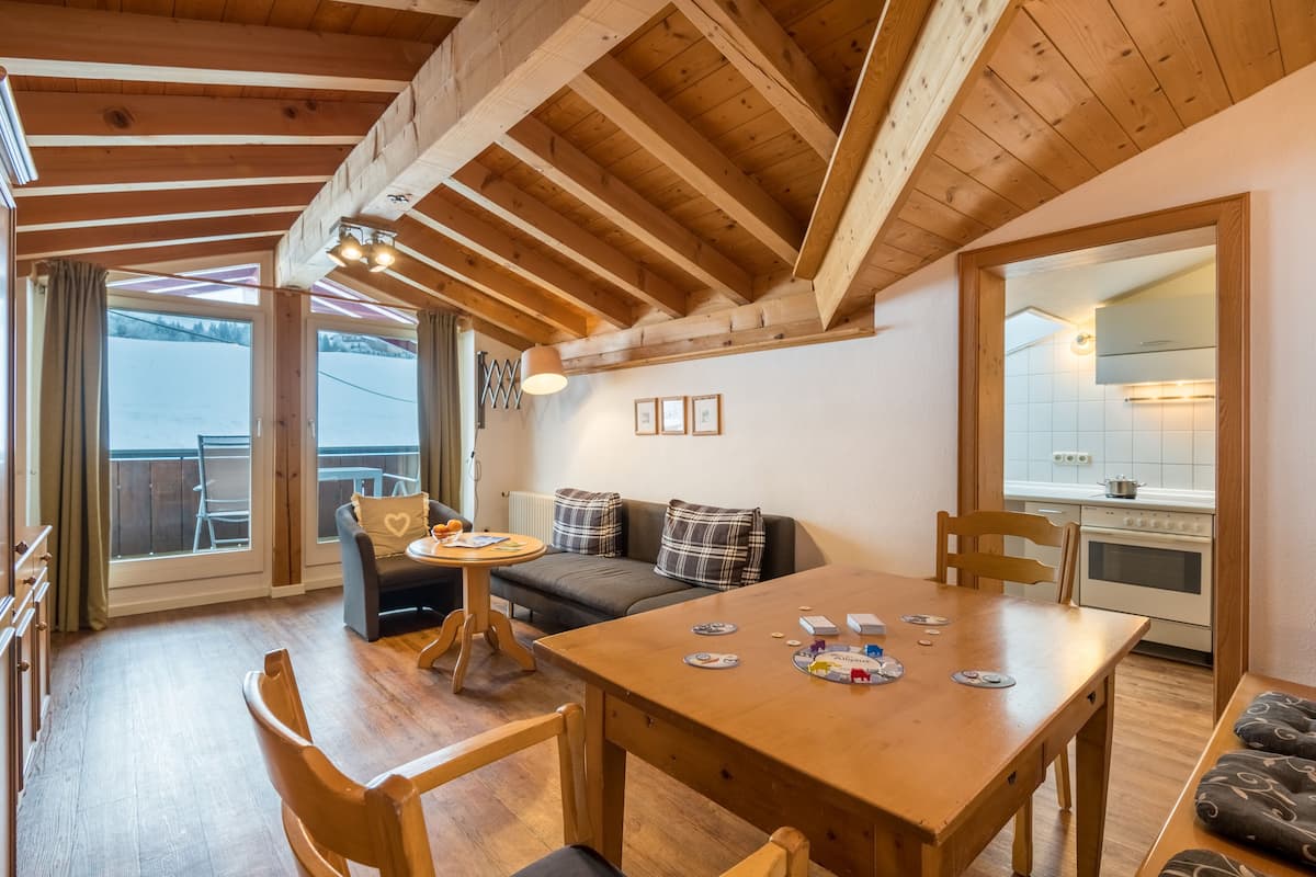 Una habitación acogedora con techo de madera, una mesa de comedor con sillas, un sofá y vistas a un paisaje nevado a través de grandes ventanales.